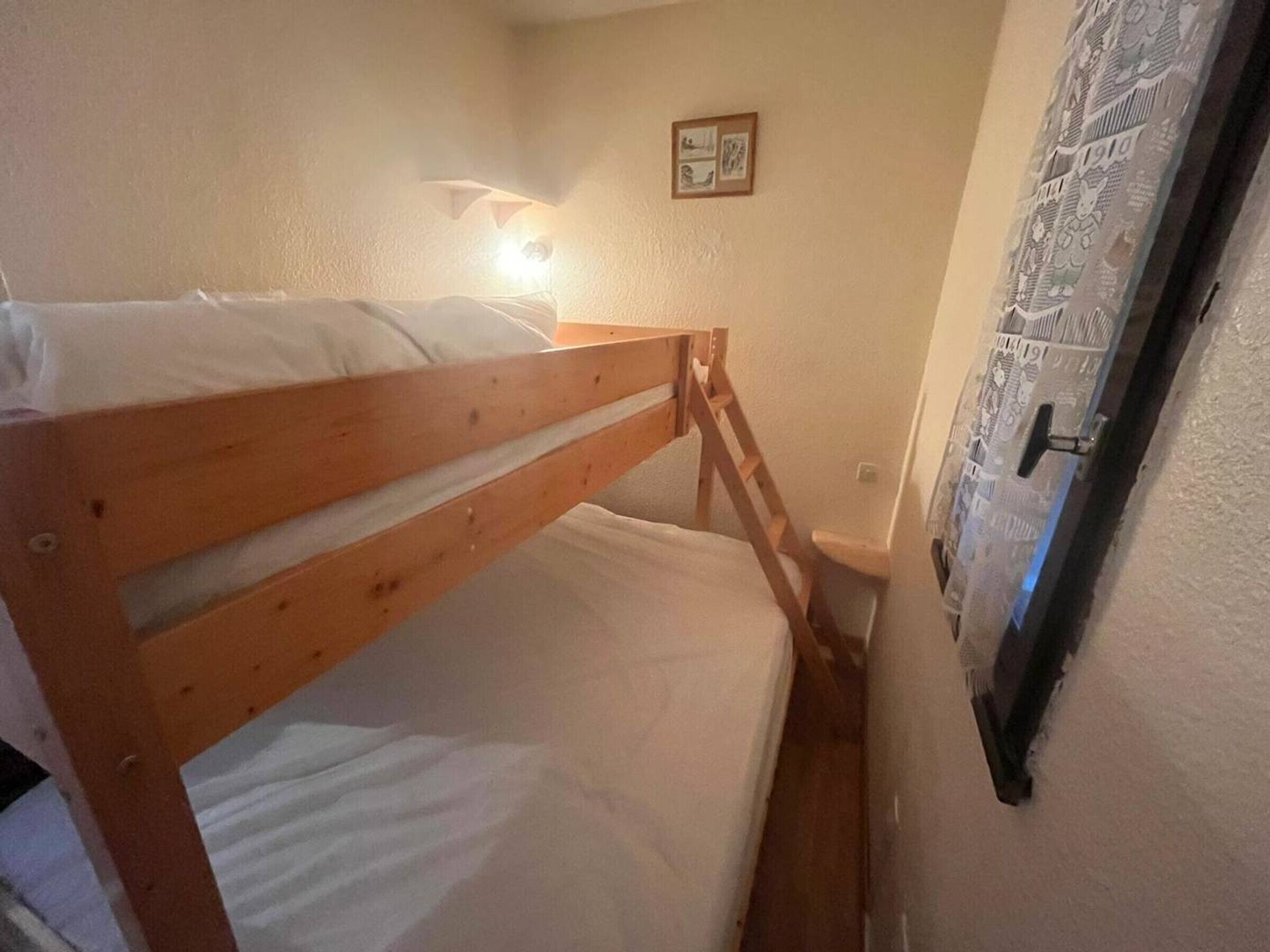 A small, narrow room with a wooden bunk bed, a wall-mounted light, and a door with a patterned curtain.