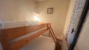 A small, narrow room with a wooden bunk bed, a wall-mounted light, and a door with a patterned curtain.