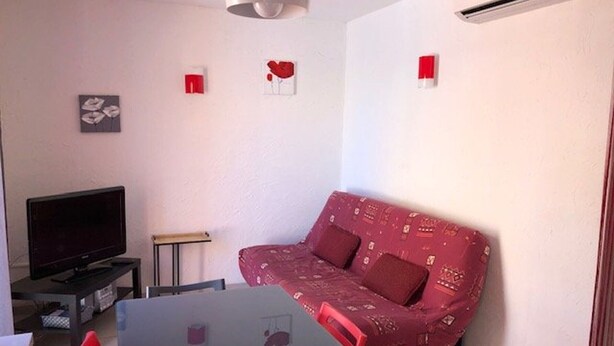 A living room with a red patterned sofa, a flat-screen TV, a table, and a ceiling light. - Quiet Air-Conditioned Apartment with Parking Near Center and Beach for 4 (Cap d'Agde)