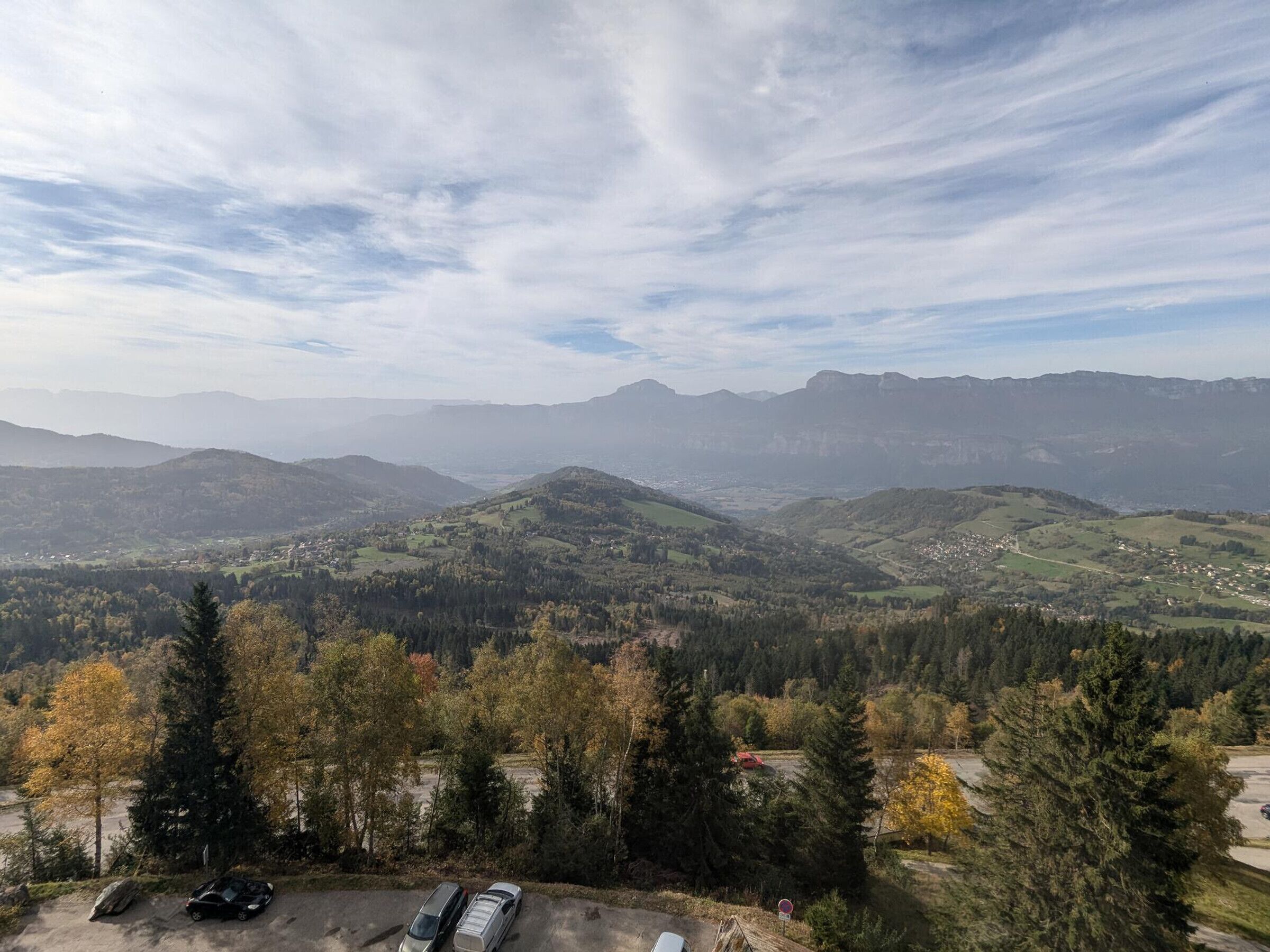 A panoramic view of a mountainous landscape with a valley below, featuring a parking area with cars and a clear sky.