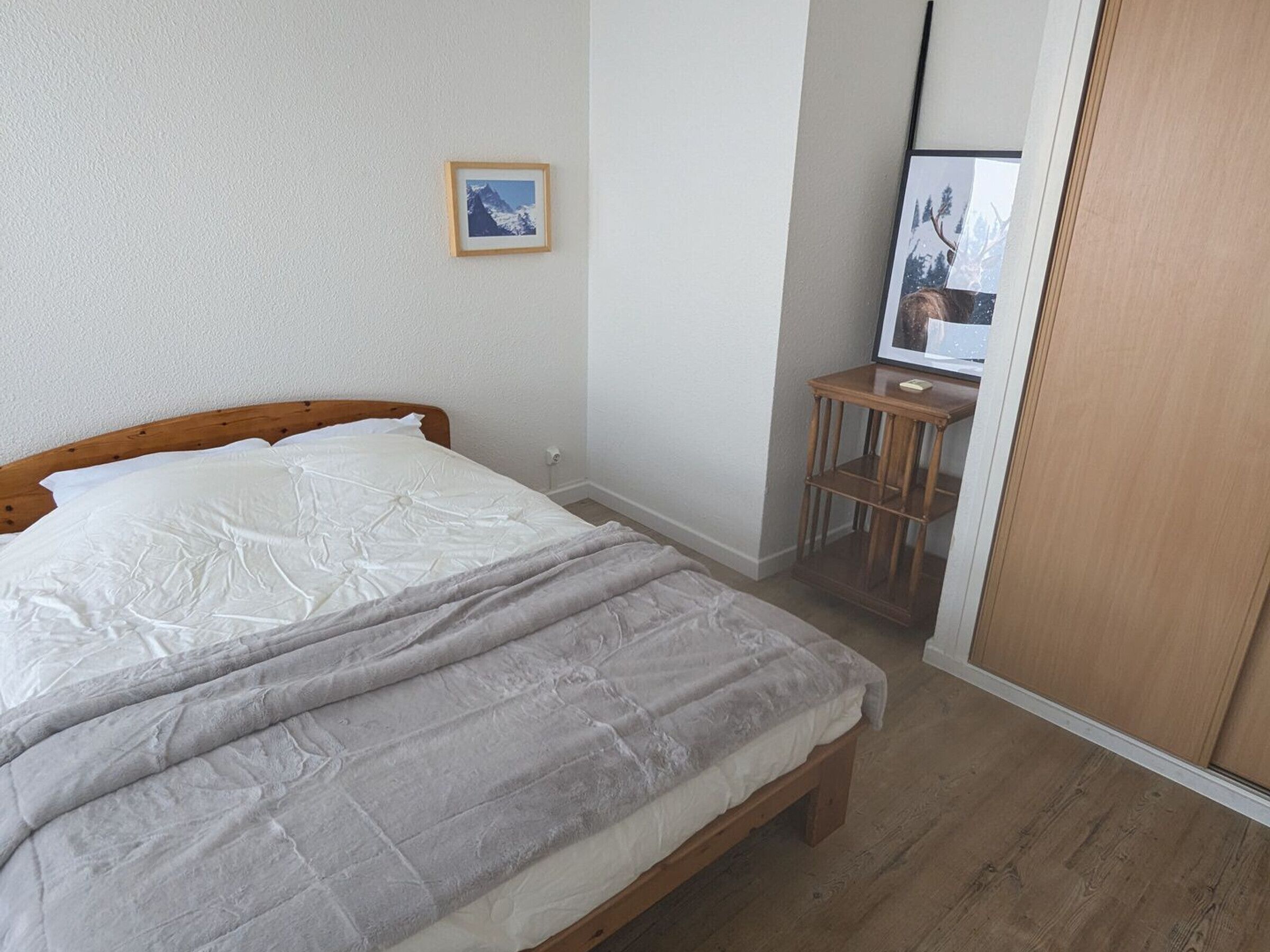 A bedroom with a wooden bed, a bedside table, a small wooden stool, and a framed picture on the wall.