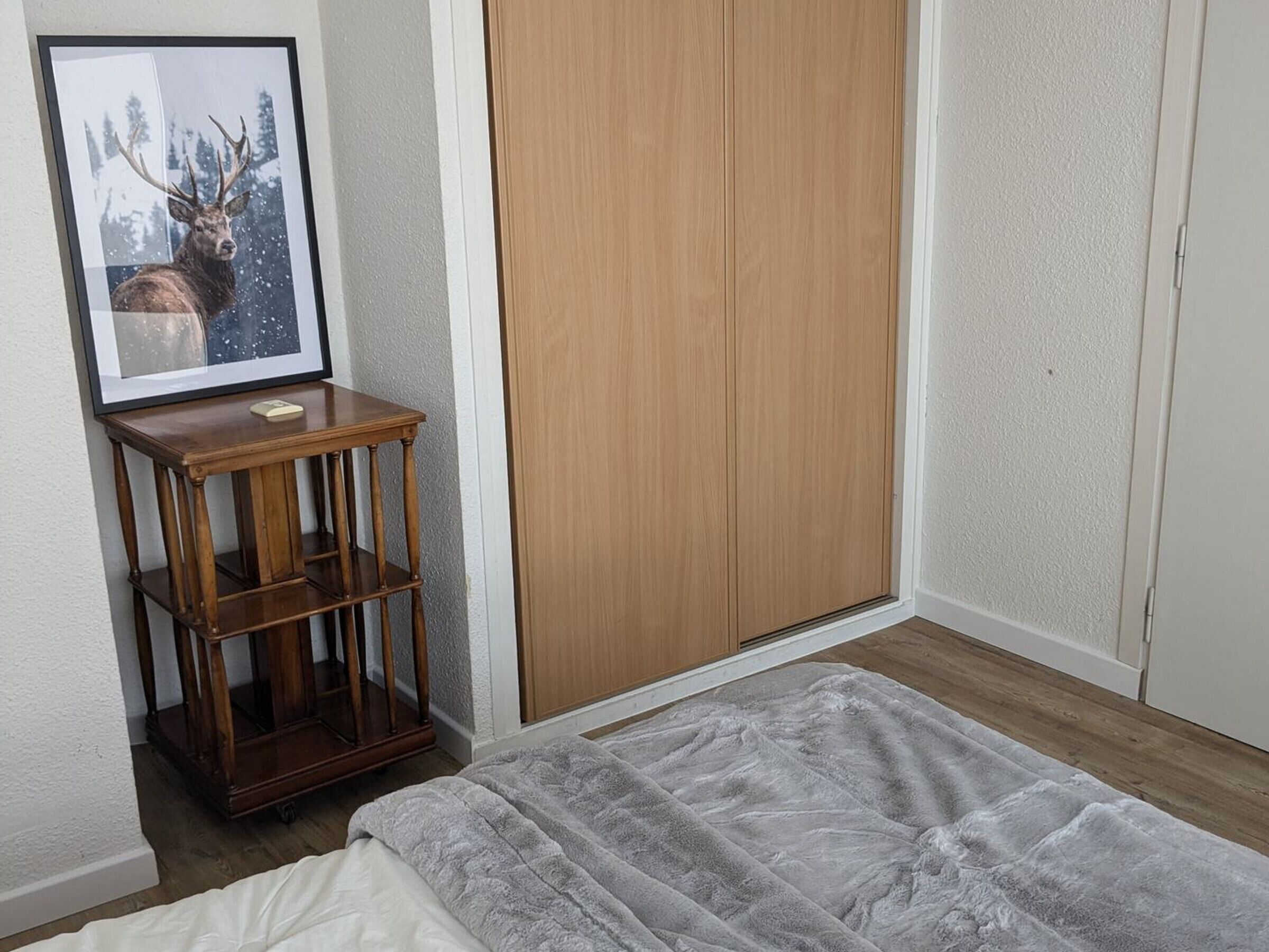 A bedroom with a bed, a wooden wardrobe, a framed picture of a deer, and a small wooden table.