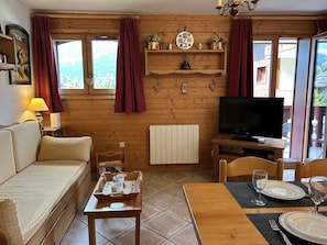 Uma cabana de madeira acolhedora com um sofá, uma mesa de jantar posta para dois, uma televisão e uma janela com cortinas.