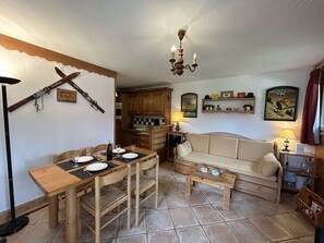Uma sala de estar acolhedora com uma mesa de jantar de madeira, sofá e mesa de centro. Uma cozinha é visível ao fundo.