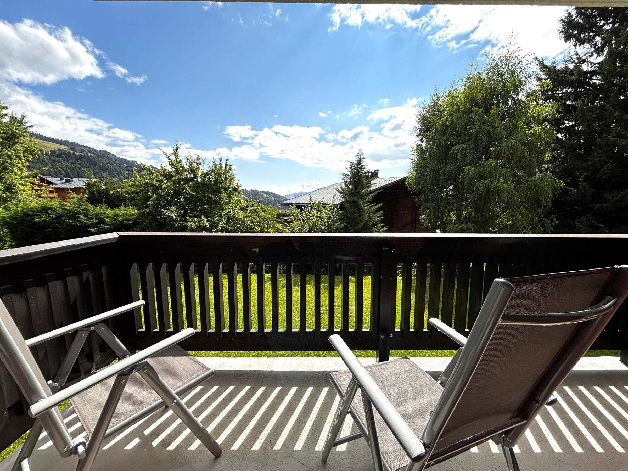 Un balcon avec deux chaises en métal et une table, donnant sur un espace verdoyant avec des arbres et une montagne en arrière-plan.