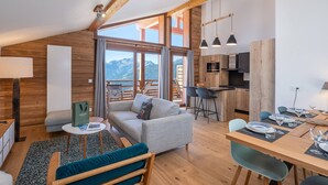 A modern interior with a loft-style ceiling, a cozy seating area, a kitchen, and a dining space with a mountain view. - Spacious duplex in Les Saisies for 8 people with parking included (Les Saisies)