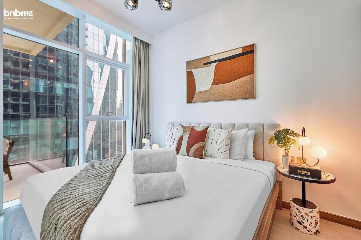 Une chambre d’hôtel moderne, dotée d’un grand lit, d’une table de chevet avec une lampe et offrant une vue sur la ville grâce à de grandes fenêtres.