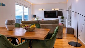 A modern living room with a wooden dining table, green upholstered chairs, a sofa, and a flat-screen TV mounted on the wall.