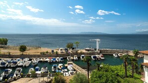 A marina with boats docked, a clear sky, and a view of the sea and distant mountains.