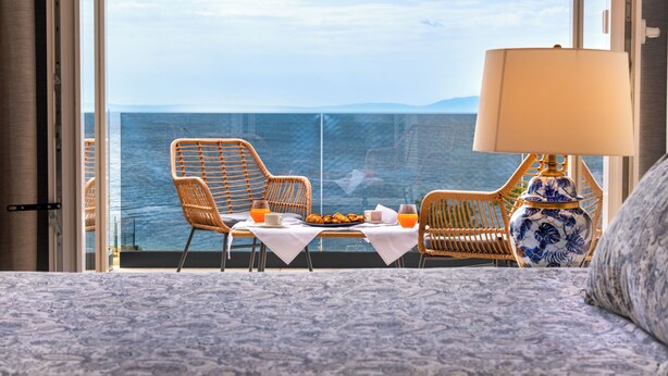 A balcony with wicker chairs, a dining table with breakfast, and a view of the sea.