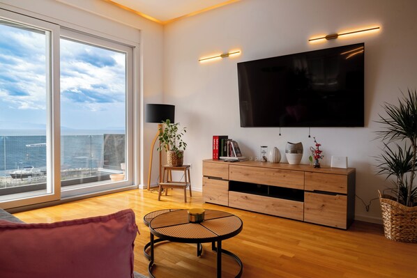 Una sala moderna con un gran ventanal, piso de madera, una mesa redonda, un televisor montado en la pared y un mueble de madera con objetos decorativos.