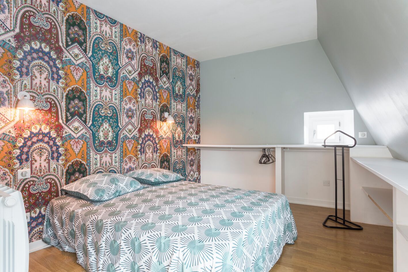 Un dormitorio con pared estampada, una cama con cubrecama de diseño floral, una mesita de noche y una lámpara colgante.