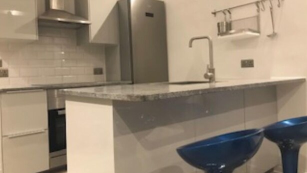 Una cucina moderna con un frigorifero in acciaio inossidabile, un forno integrato e un lavello con rubinetto. Sono presenti due sgabelli da bar blu e un portaspezie a sospensione sopra il lavello.