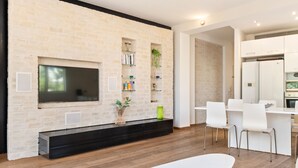 Una sala moderna con un televisor de pantalla plana montado en una pared de ladrillo, un banco de madera y una zona de comedor con sillas y una mesa blancos.