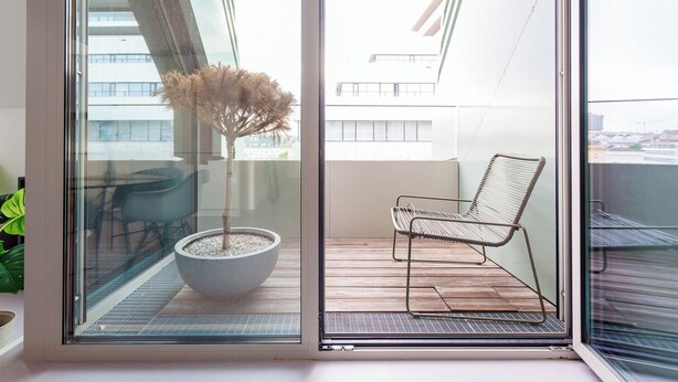A balcony with a potted tree, a metal chair, and a view of buildings through a glass door. - JULIUS 23: Cozy Stay - 5 Mins to City Center (Wien)