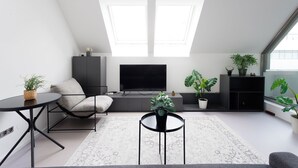A modern loft apartment with a large TV, two skylights, a sofa, a coffee table, and several potted plants. - JULIUS 23: Cozy Stay - 5 Mins to City Center (Wien)