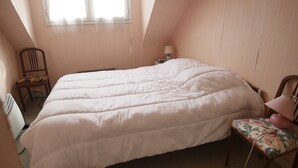 Un dormitorio con una cama grande, una silla, una mesita de noche con lámpara y una ventana con cortinas.