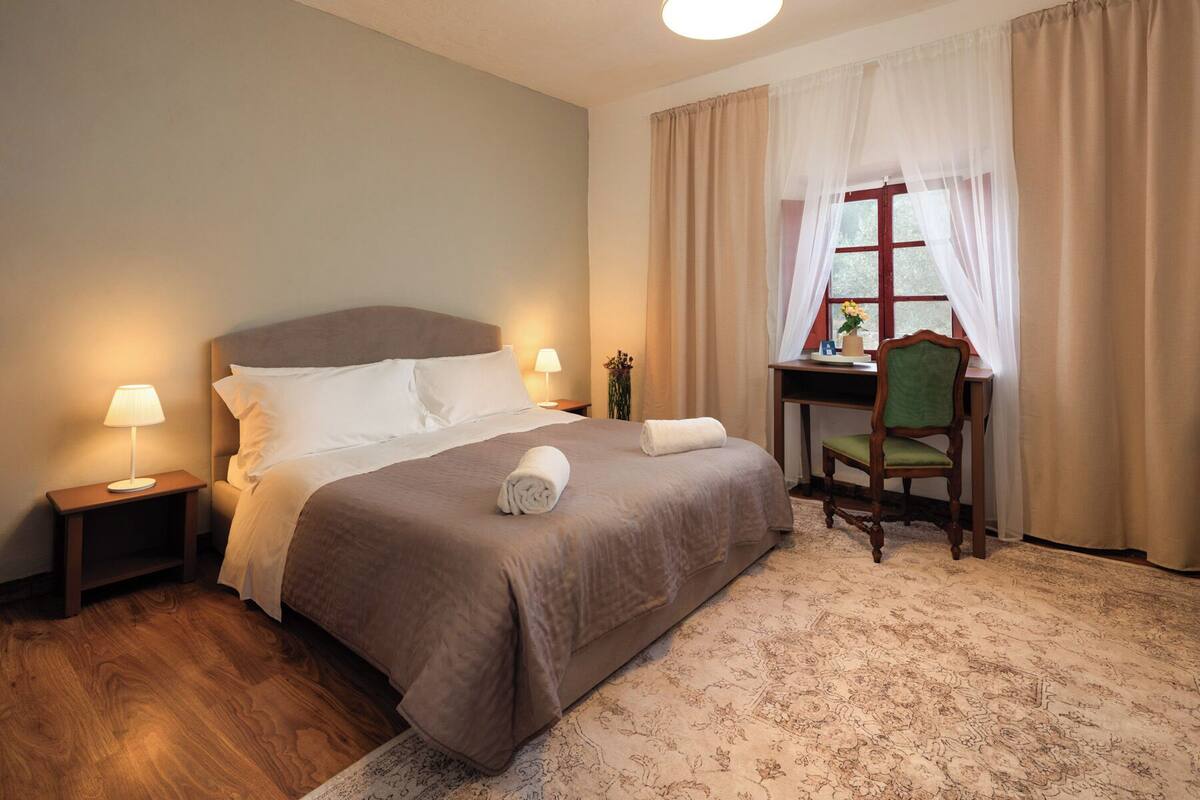 Habitación de hotel con cama, lámparas de noche, escritorio con silla, ventana con cortinas y alfombra.