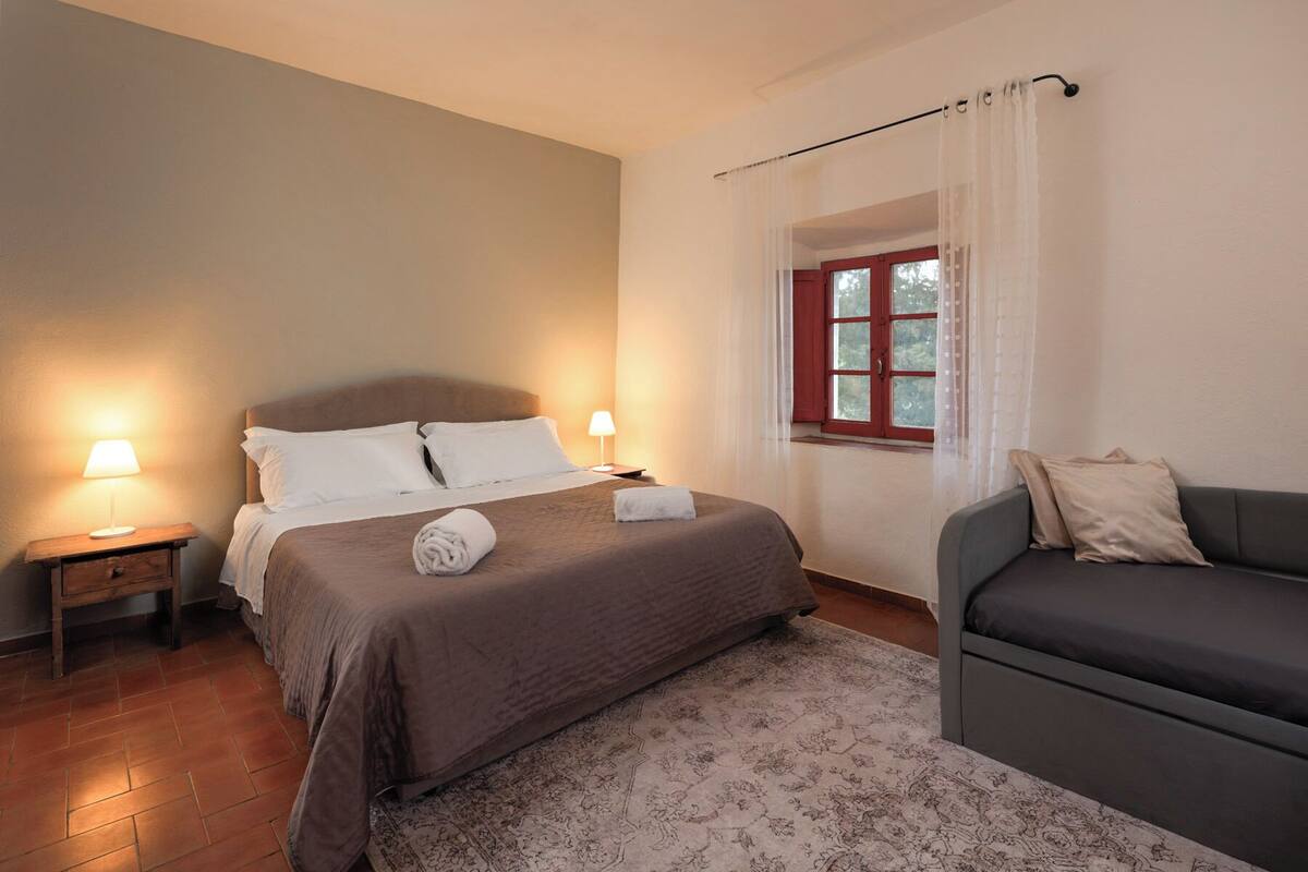 Un dormitorio con una cama, lámparas de noche, una ventana con cortinas, un sofá y una alfombra estampada.