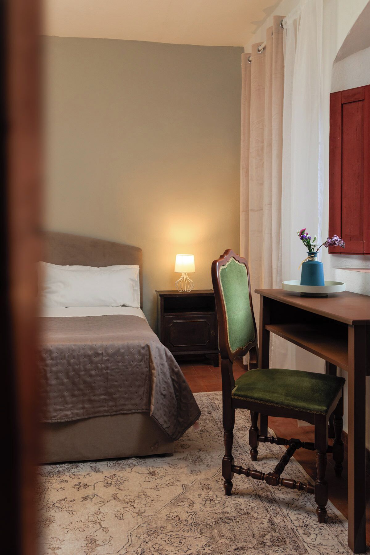 A hotel room with a bed, a desk with a chair, a lamp, and a vase with flowers.