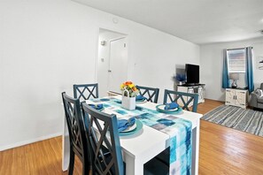 Een eetruimte met een tafel gedekt voor vier personen, stoelen, een vaas met bloemen, een televisie op een standaard en een kast.