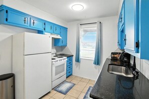 A compact kitchen with white appliances, blue cabinets, and a window with curtains.