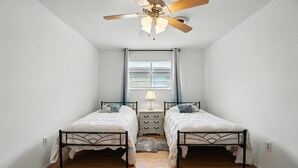 Een slaapkamer met twee eenpersoonsbedden, een plafondventilator, een raam met zonwering en een nachtkastje met een lamp.