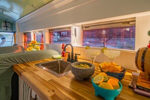 Een keukenhoek met een spoelbak, een stenen vijzel, een fruitschaal en een raam met gordijnen.
