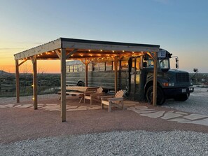 Een overdekt terras met houten stoelen en een bankje, een bus, en een grindpad.
