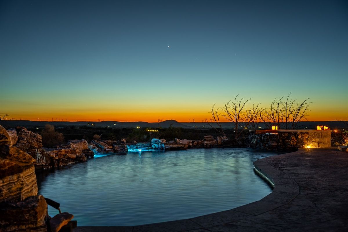 A nighttime view of a swimming pool with a waterfall feature, surrounded by rocks and a clear sky.