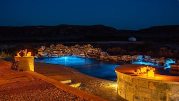 Une piscine extérieure éclairée pour la baignade nocturne, agrémentée d’une cascade en pierre, entourée d’une terrasse et d’un mur en pierre.