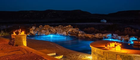 A nighttime outdoor swimming pool with a stone waterfall feature, surrounded by a stone deck and a stone wall.