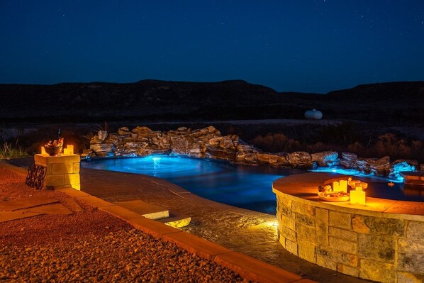 A nighttime outdoor swimming pool with a stone waterfall feature, surrounded by a stone deck and a stone wall.