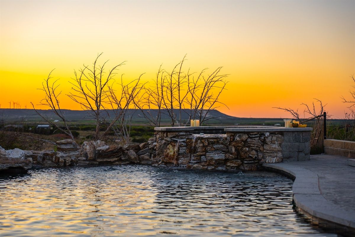 A stone water feature with a stone bench and candles, surrounded by a landscape with trees and a sunset.