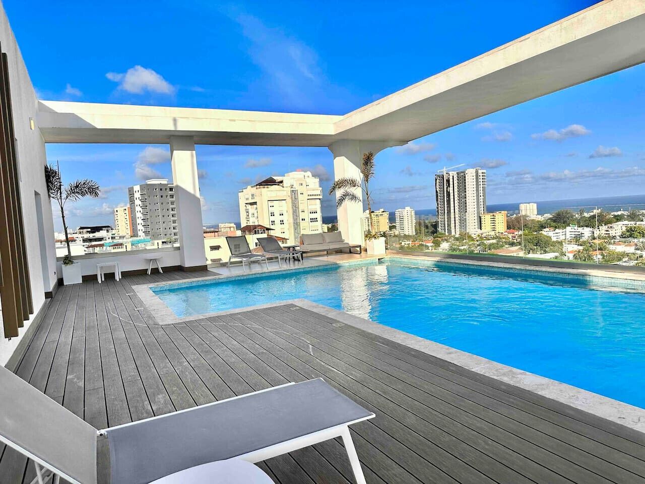 Un espace piscine aménagé sur le toit, avec une terrasse en bois, des chaises longues et une vue sur le paysage urbain.
