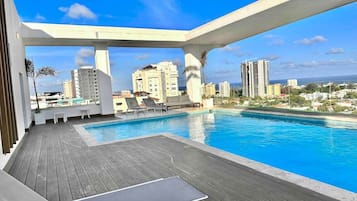 Un espace piscine aménagé sur le toit, avec une terrasse en bois, des chaises longues et une vue sur le paysage urbain.