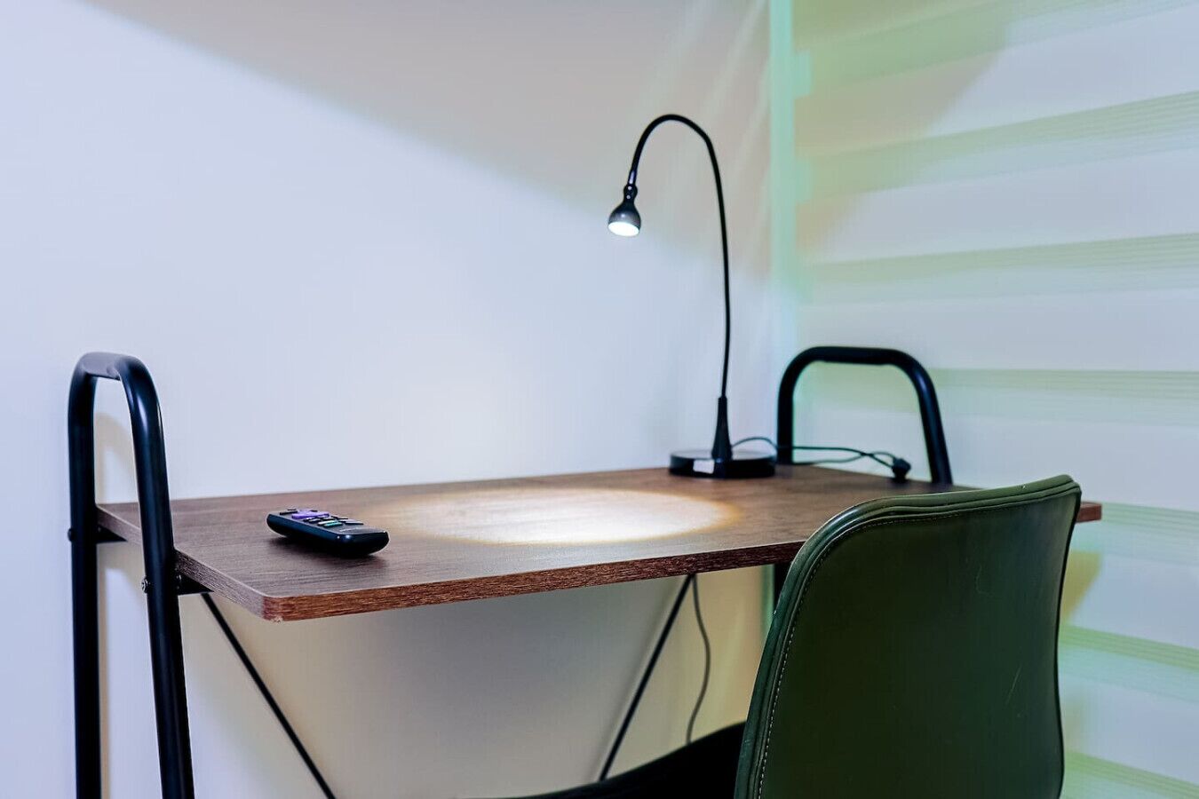 Un petit bureau en bois, avec une lampe, une télécommande et une chaise verte.