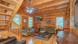 A cozy log cabin interior with a wooden staircase, two sofas, a coffee table, and a TV mounted on the wall.