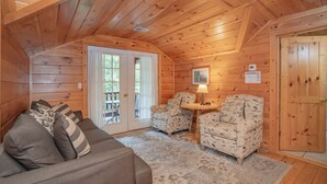 A cozy cabin interior with a wooden ceiling, a sofa with patterned cushions, two patterned armchairs, a small table with a lamp, and a door with a wooden panel.