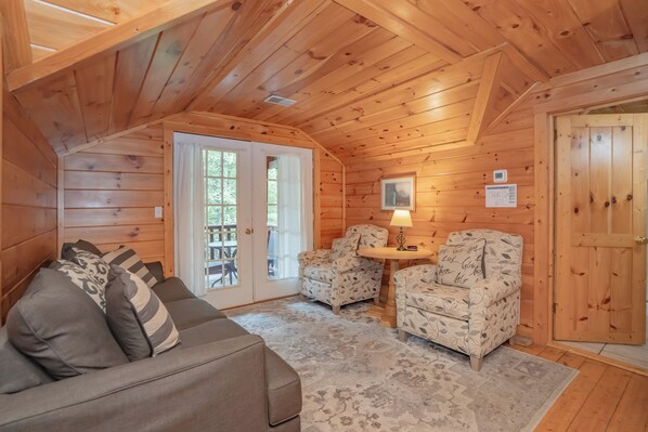 A cozy cabin interior with a wooden ceiling, a sofa with patterned cushions, two patterned armchairs, a small table with a lamp, and a door with a wooden panel.