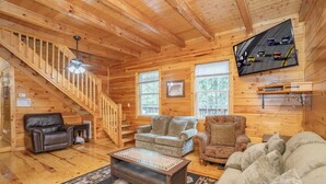 A cozy log cabin interior with a staircase, two sofas, a coffee table, and a TV displaying a road scene.