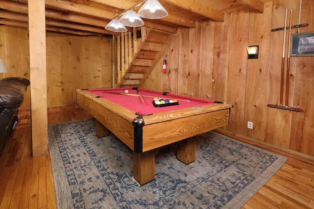 Une table de billard en bois, recouverte d’un drap de feutre rouge, est entourée d’un tapis à motifs, dans une pièce aux murs en bois et dotée d’un escalier.