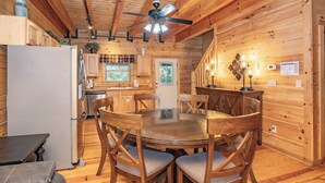 L’intérieur d’une cabane en bois comprend une table de salle à manger ronde, des chaises en bois, un ventilateur de plafond et un réfrigérateur.
