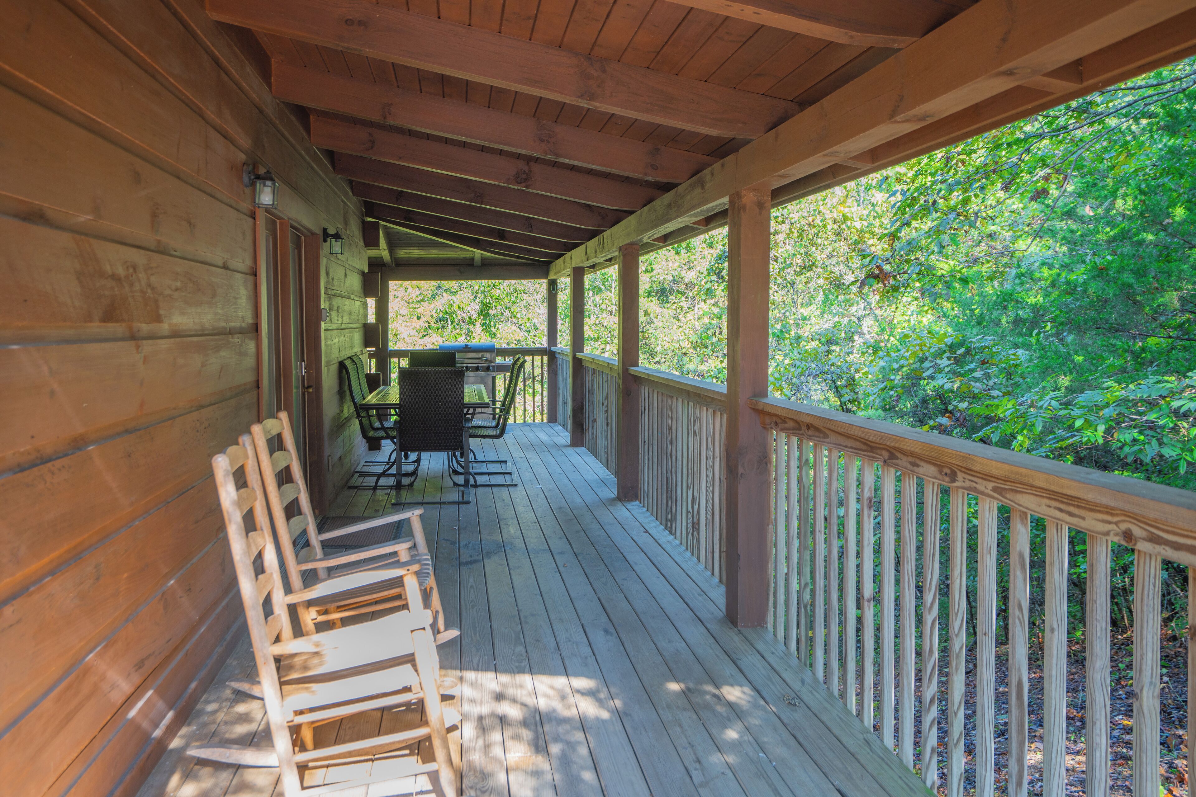 A wooden deck with a table and chairs, surrounded by a railing, overlooking a forested area.