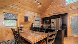 A wooden cabin interior with a dining table, chairs, a kitchen area, and a large window with patterned curtains. - 🌳 Keaton's Cozy Cabin 🌲 Wooded Retreat, Resort Pool & Hot Tub (Sevierville)