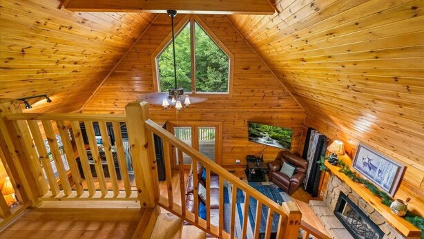 A wooden cabin interior with a loft bedroom, a fireplace, and a large window overlooking greenery. - 🌳 Keaton's Cozy Cabin 🌲 Wooded Retreat, Resort Pool & Hot Tub (Sevierville)