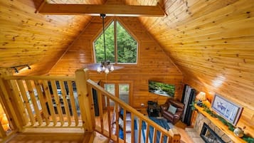 A wooden cabin interior with a loft bedroom, a fireplace, and a large window overlooking greenery.