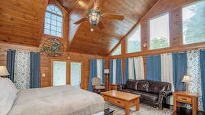 A wooden cabin interior with a bed, a leather sofa, a coffee table, and a ceiling fan. - Keaton's Firefly Meadow: OutdoorPool*HotTub*Games*Wifi*FirePit*Yard*Large Groups (Sevierville)