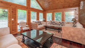A cozy cabin interior with wooden walls, large windows, and a mix of seating areas including a sofa, chairs, and a coffee table. - Keaton's Firefly Meadow: OutdoorPool*HotTub*Games*Wifi*FirePit*Yard*Large Groups (Sevierville)