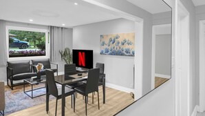 Ett modernt vardagsrum med en matplats, ett stort fönster med utsikt över en bil och blommor, en platt-TV och en tavla på väggen.